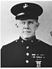 Head and shoulders of a white man wearing a peaked cap and a dark military jacket with ribbon bars on the left breast and bright buttons down the center.