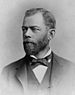 Head and shoulders of a white man with a full beard, wearing a dark suit coat, vest, and bow tie.