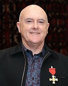 Murray looks at the camera. He has a bald head, grey eybows and is wearing a paisley shirt and a dark jacket the displays his New Years award medal. He is smiling.