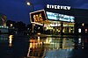 A bright marquee is reflected over a wet, dark street; the name Riverview is visible above.