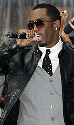 A man wearing sunglasses and a waistcoat with a leather jacket on top singles into a microphone