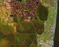 A color satellite image of the northern Everglades showing green chunks of Everglades surrounded by white settlement areas of the South Florida Metropolitan Area to the east and red agricultural fields in the Everglades Agricultural Area to the north