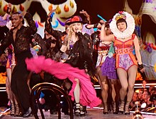 Picture of a group of people dressed in colorful, Mexican-styled clothing. The woman in the middle is blonde, wears a black hat with a pink flower, and waves her pink skirt towards the camera. Her hair is braided and she sings to a microphone in her mouth. To her right, there is a female in an orange and purple leotard, and a white wreath around her head. Also present is a black male in dark clothing.