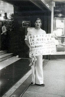A man with a sign and a plastic bag in front of a mall door.