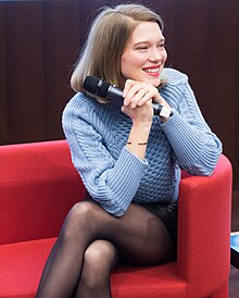 Photographie au format portrait et en couleurs d'une femme assise sur le fauteuil d'une scène de conférence, un micro à la main.