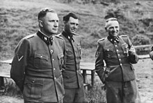 Three men in SS uniform stand outdoors looking at viewer's right.