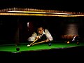 A man playing snooker on a baize-covered table, preparing to strike the white ball using a cue stick that he is holding with his right hand; the end of the cue stick is resting on the cross-shaped head of a long stick which he is holding with his left hand