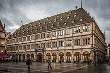 Photographie d'un bâtiment avec des arcades, deux étages et un grand toit percé par des lucarnes.