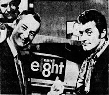 Bob Wells and Chuck Schodowski pose in front of a television camera with a large "ei8ht" logo on it. The i in "ei8ht" is dotted with the CBS eye, and above the 8 in small text reads "WJW-TV".