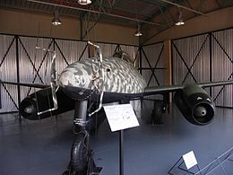 A twin engine jet aircraft pictured from front-left sitting on the ground in a hangar. The paint scheme of the aircraft is camouflage of various brown and green colors. Two antennas are protruding from the nose of the aircraft. The white number "305" is visible on the nose of the aircraft.