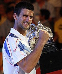 A brown-haired man in a white tennis shirt with light blue sections and red stripes with the trophy