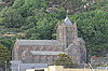 A church seen from the south at a distance with a tree-covered rocky hillside beyond; the church is large with a clerestory, a castellated tower with a small pyramidal spire and clock faces on the two visible sides