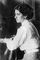 A Caucasian female sitting at a desk holding a pen, with her face in profile. She is wearing a white high collared long sleeved shirt, belt, and checkered bottom with dark upswept hair.