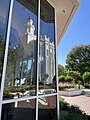 A glass window from the visitors center reflects the St. George Temple, with tall white parapets and a domed octagonal spire. Various foliage is seen off to the side.