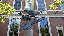 Two Deinonychus figures balance on a claw-like form, in active poses as if they are pursuing prey. In the background is a banner advertising the The Academy of Natural Sciences, and the brick structure and windows of the museum itself. It's late in the afternoon on a sunny day in July, and a sunbeam striking the face of one of the sculpted dinosaurs casts dramatic shadows.