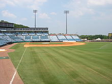 A view from right field shows the green grass and infield dirt of a baseball diamond surrounded by empty blue seats