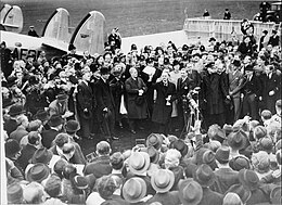 A large crowd on an airfield; British prime minister Neville Chamberlain presents an assurance from German chancellor Adolf Hitler.