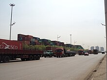 line of heavy trucks going north, background a container yard