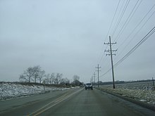 Westbound IL 120 just west of city of McHenry and narrowing to two lanes. Riding on concrete, there are open areas on both sides of the road with scattered trees on the left and utility poles on the right.