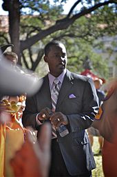 Desmond Howard wearing a grey striped suit while holding a water bottle outside. He has his XXXI Super Bowl ring on his right ring finger and his wedding ring on his left ring finger.