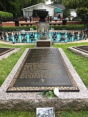 A long, ground-level gravestone reads "Elvis Aaron Presley", followed by the singer's dates, the names of his parents and daughter, and several paragraphs of smaller text. In the background is a small round pool, with a low decorative metal fence and several fountains.
