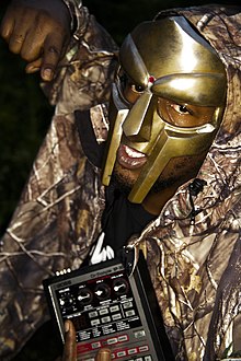 Photo portrait of a man wearing a golden mask and hoodie, holding a sampler and pointing at the viewer