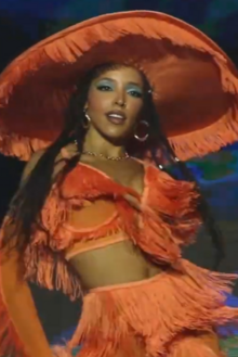 A woman wearing a showy orange costume and big hat dances.