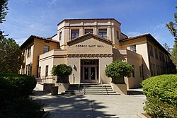 The entrance of a light colored Spanish revival building with steps leading to a double door in sunlight. Bushes surround the outside with some trees in the far background.