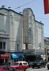 Color photograph of the exterior of a movie theater, showing 1990s-2000s-era automobiles on the street in front of the theater, electric lines above the cars, and shop windows and awnings along the sidewalk. The theater features a large vertical 'blade' that reads "Castro", a marquee announcing film showings, and a facade which includes a mission-revival arch and highly ornamented panels designed in the Churrigueresque style.