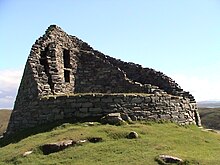 The ruins of a double-walled circular stone tower on top of a green hillock with a blue sky in the background