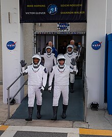 3 male astronauts and one female astronaut all in white spacesuits smile and wave their hands while walking down the ramp of a building adorned with NASA logos and mission patches