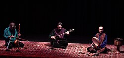 Kayhan Kalhor, Hossein Alizâdeh, and Majid Khalaj sit on a long Persian rug on a dimly-lit stage. The heads of Kalhor and Alizâdeh are lowered in concentration over their stringed instruments. Khalaj has closed eyes and a countenance of calm focus as his fingers strum the frame drum in his hand.