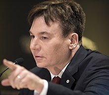 A close up photo of Mike Myers wearing a black blazer with a red tie. He appears to be pointing at something.