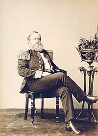 Photograph of a bearded man seated casually with crossed legs and wearing a military tunic with fringed epaulettes