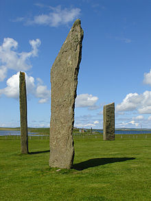 Three tall thin monoliths sit in a grassy field under blue skies with two bodies of water beyond at right and left.