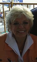 A woman with short blond hair, wearing a white shirt with an orange sweater.