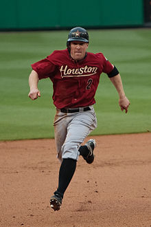 A white man in a red baseball uniform running bases. Looking into the camera, he has on a red baseball uniform reading "Houston" in yellow text with similarly colored line under it. Below that to the runner's left hand side, there is a black "2" with a red outline. In addition to a black baseball with an orange star, he has on gray pants, black socks, and black cleats.