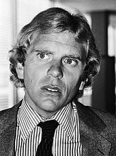 A black and white photograph of a middle-aged man of British appearance. The man is clean shaven and sports curly hair. He is wearing formal attire, a striped shirt with a black tie is visible under a grey suit jacket. He is looking past the viewers left.