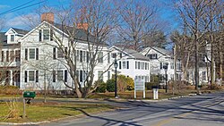 A row of two-story white houses with black or gray gabled roofs down a street on the left of the image. In the rear, slightly obscured by some bare trees, is a similar church with a short four-pointed steeple.