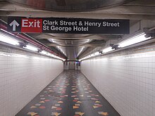 A passageway above the platforms, leading to the elevators. The passageway has black floors with multicolored shapes, which are part of an artwork. The walls are clad with white tiles. There is a sign on the ceiling, directing passengers to the station's exit in the St. George Hotel.