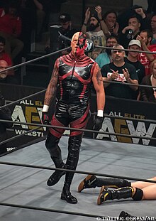 A color photograph of a man in a professional wrestling ring with a red outfit and half of his face painted red