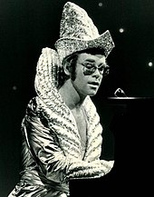 A young man wearing an elaborate shiny costume with a high collar and pointed hat playing a piano