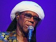 Picture of an African American man wearing glasses and a white beret; he stands in front of a purple background and speaks to a microphone.