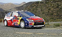 Citroën C4 WRC de Sébastien Loeb, livrée rouge, bleue et blanche, empruntant un virage serré sur une route goudronnée ; paysage montagneux en arrière-plan.