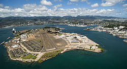 A color photo of Ford Island with modern and historical buildings surrounding a runway overrun with grass