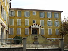 Façade ocre jaune, à deux étages et fenêtres encadrées de blanc. Fronton classique au centre et double escalier d'accès à la porte.