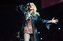 A blonde woman wearing a black leather jacket sings to a microphone.