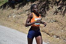 Cheptegei running along a tarmac road in an arid landscape, wearing an orange vest and dark blue shorts