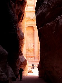 Photographie de la sortie d'un défilé de roche rouge au delà de laquelle on aperçoit la façade d'un palais sculpté dans la roche. La silhouette d'un touriste prenant une photographie à l'aide de son portable est visible à l'entrée du défilé. À l'extérieur du défilé devant le palais, se promènent deux adultes et deux enfants. Les enfants donnent la main à l'un des adultes. Un chameau se repose.