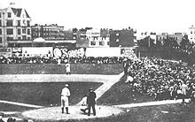 Wide shot of a black-and-white photograph of a baseball field, with spectators in the foreground and background.